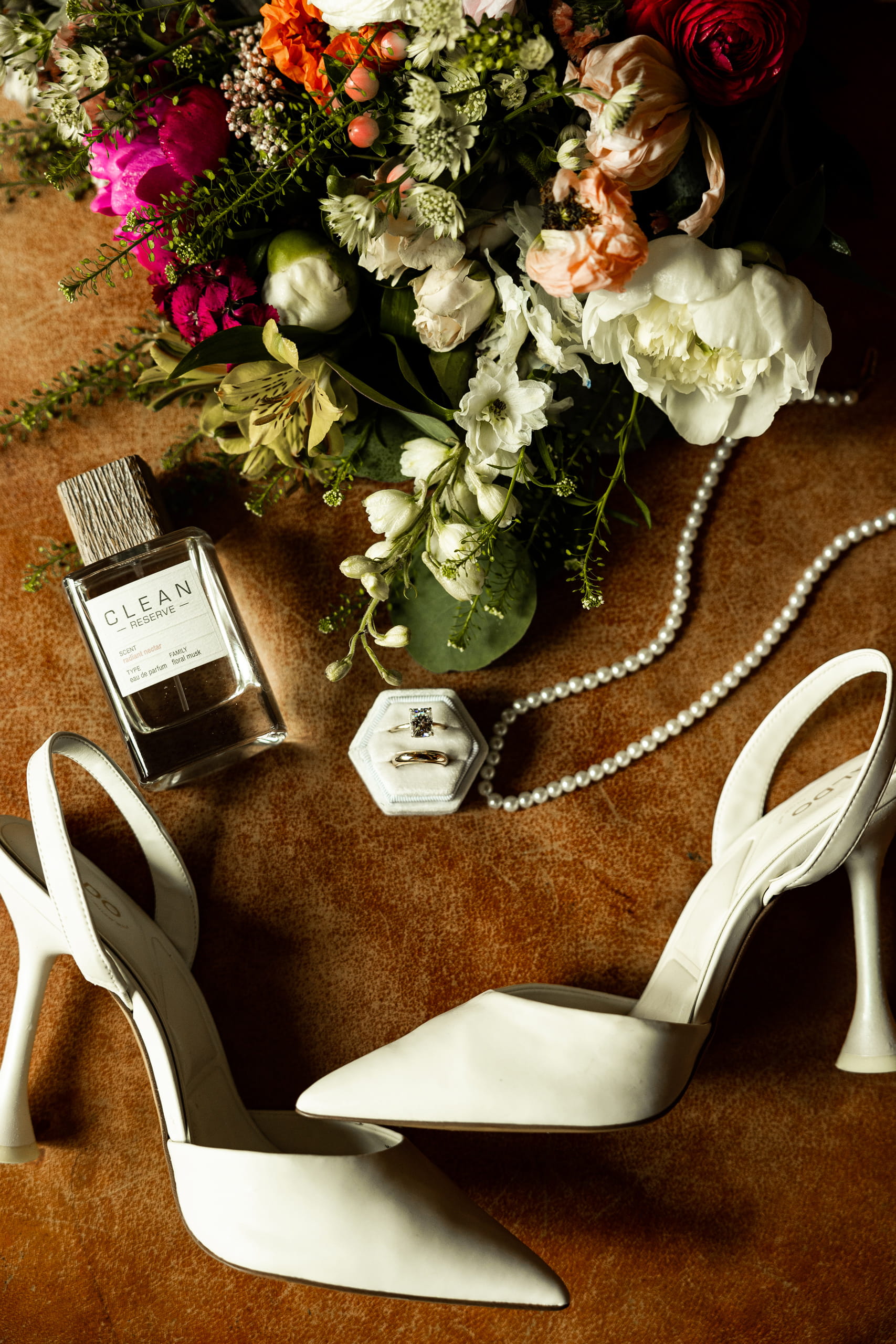 Flat lay of bridal details including bouquet, pearl necklace, perfume, shoes, and wedding rings on a brown leather background