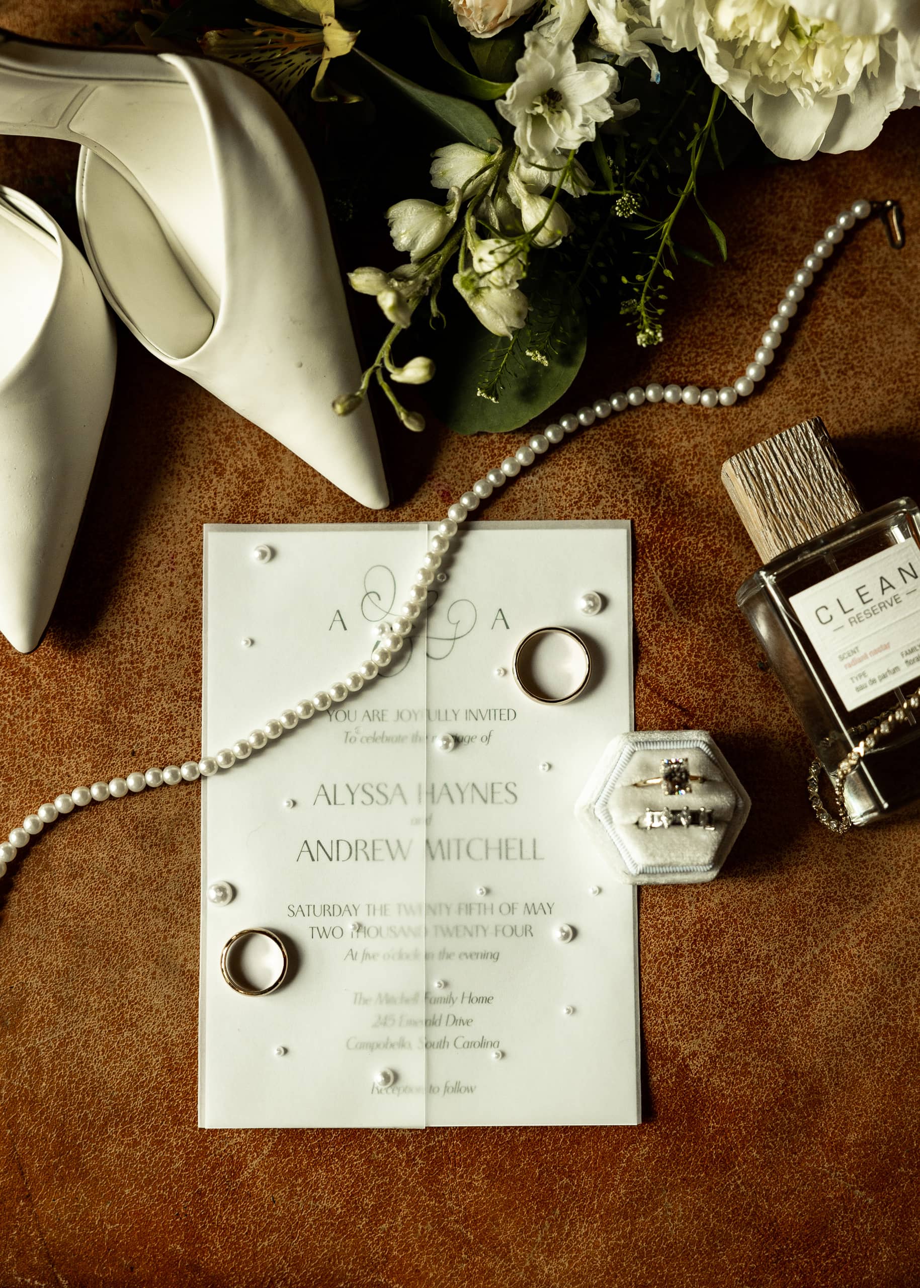 Elegant wedding flat lay with bouquet, pearl necklace, shoes, rings, perfume, bracelet and invitation arranged together on a warm brown leather surface