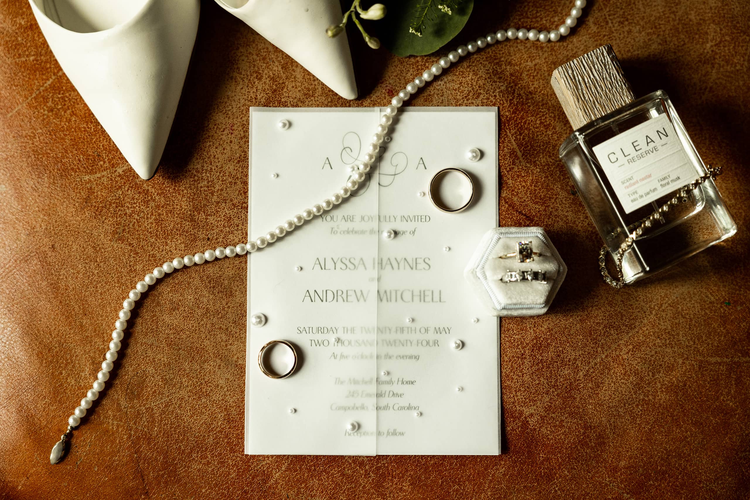 Artistic composition of all bridal details — bracelet, pearl necklace, shoes, rings, perfume, and invitation — captured from above on a brown leather background