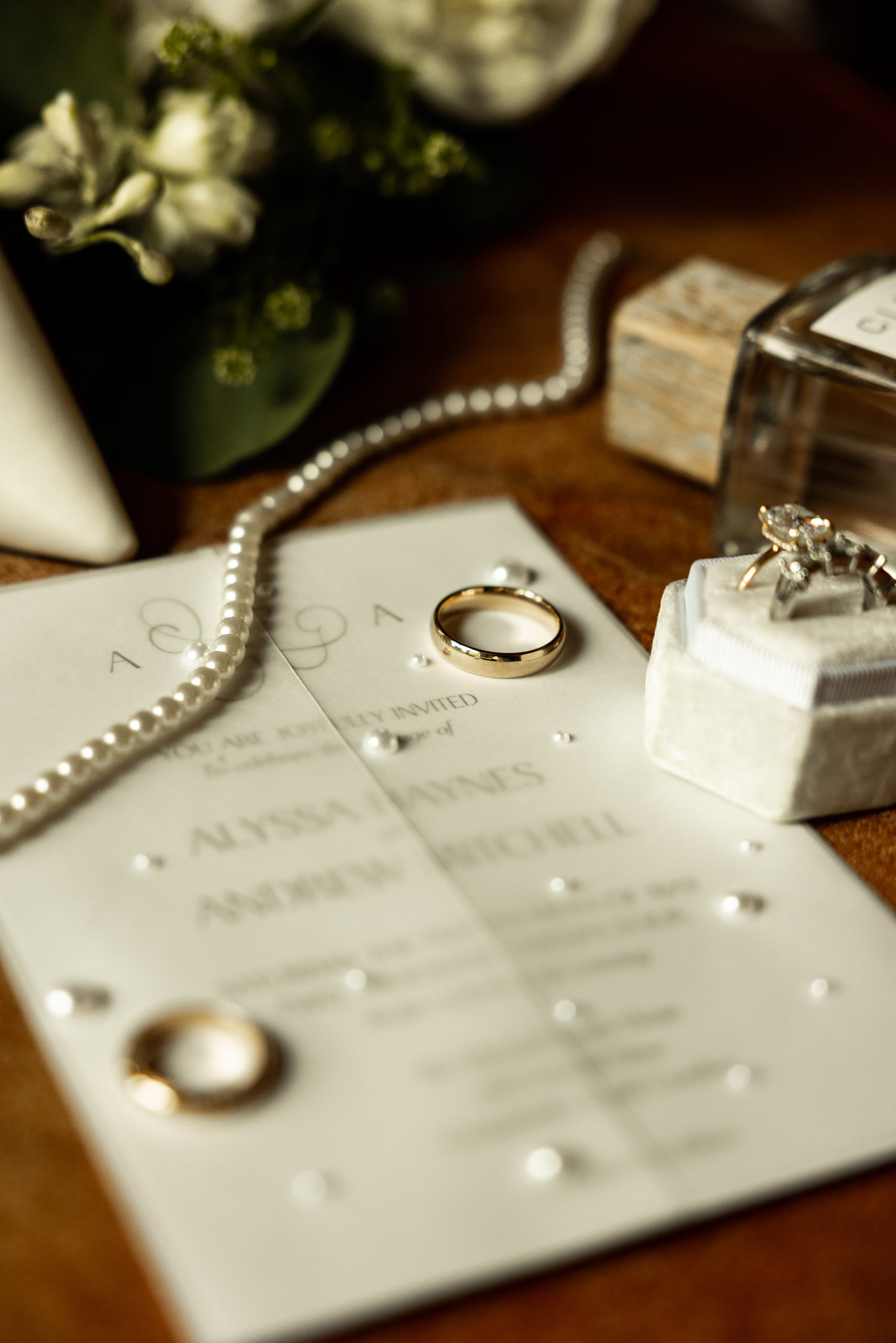 Close-up composition focusing on the wedding rings, with soft details of the bouquet, perfume, invitation, and other bridal elements on a warm brown leather surface.