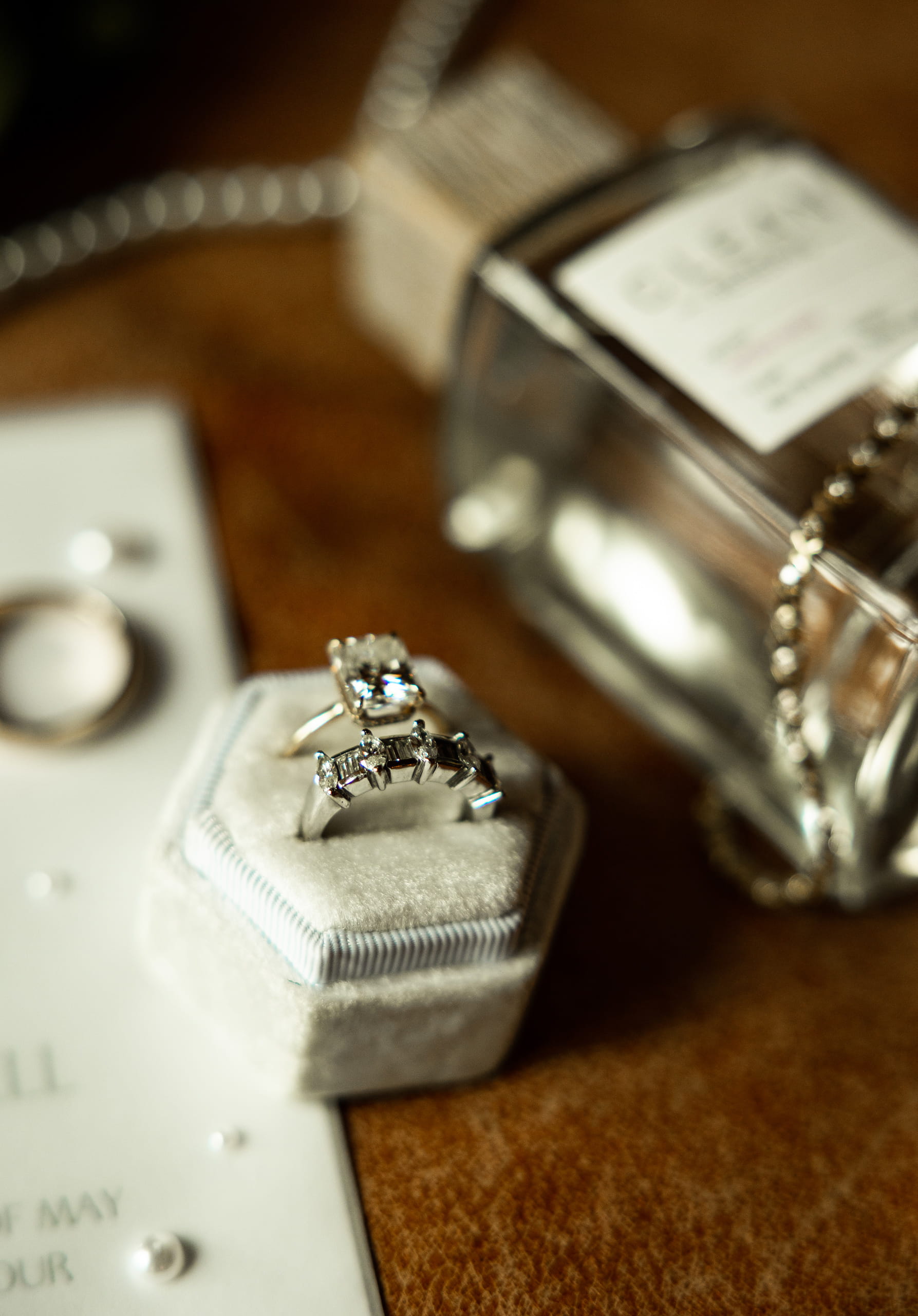Wedding rings in clear focus on a brown leather background, with the invitation, perfume, and bracelet softly out of focus behind them.