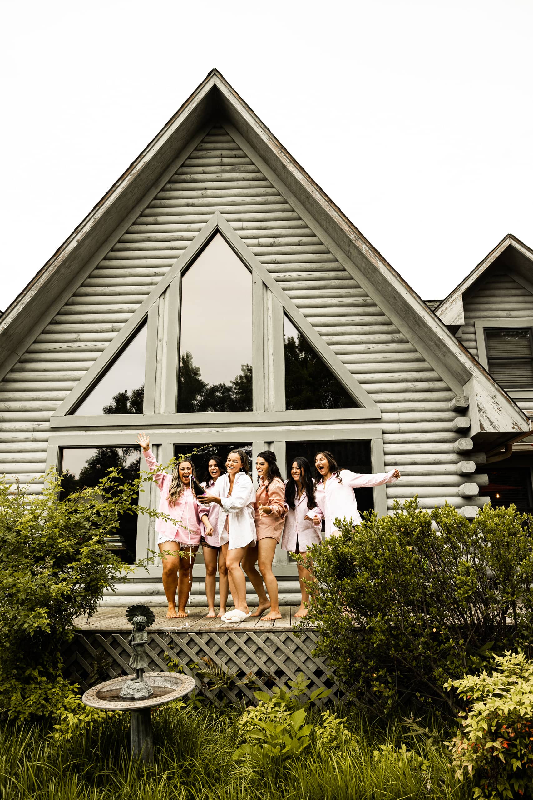 Bride and bridesmaids in matching shirts celebrating in front of the house by opening a bottle of champagne before getting ready for the ceremony.
