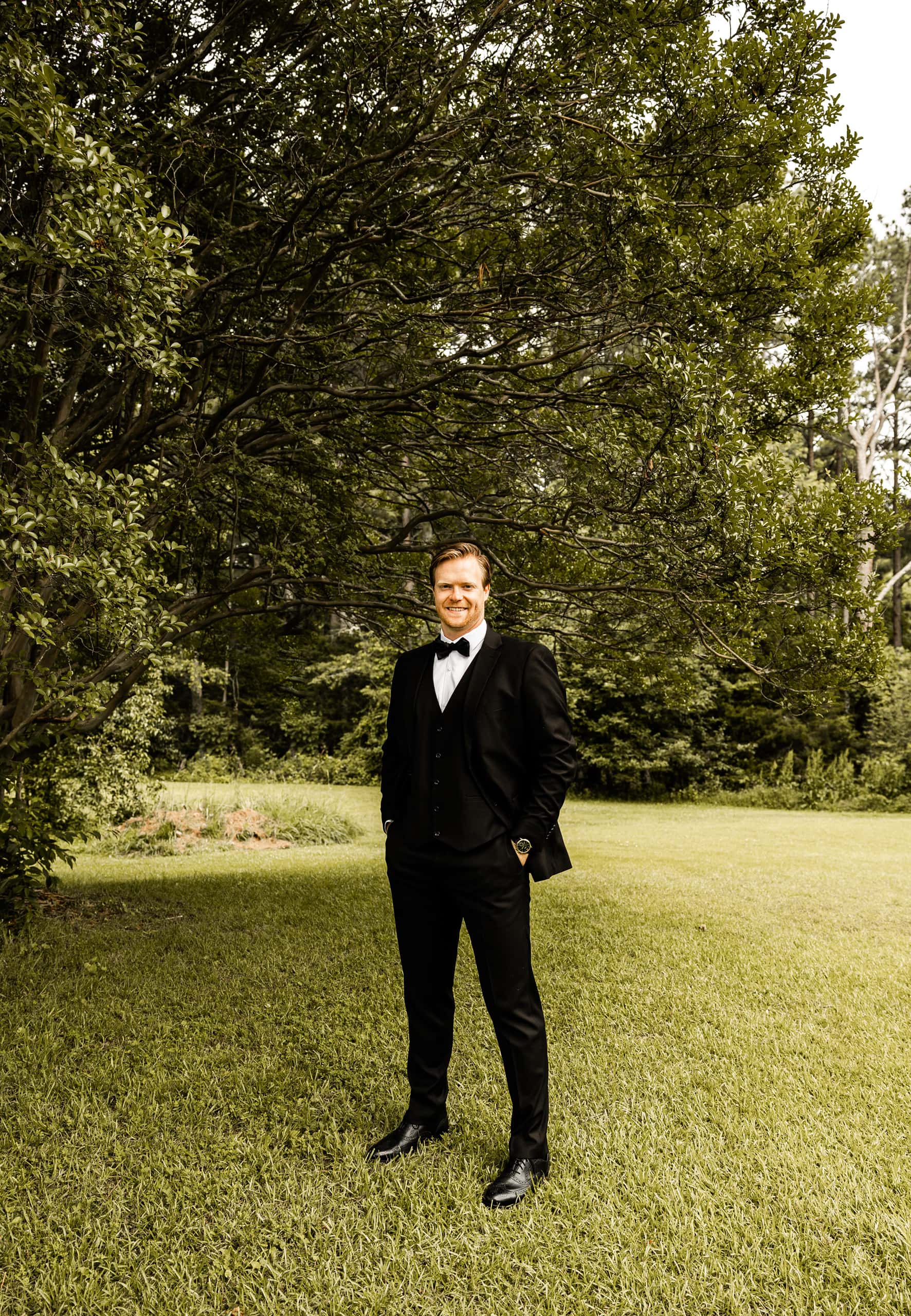 Candid image of Andrew dressed in his wedding suit, standing on the grass in the backyard, surrounded by trees and soft natural light.