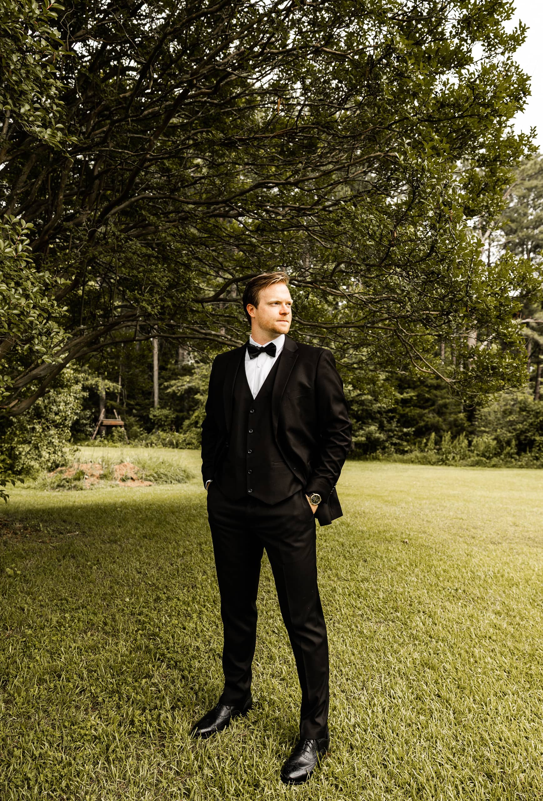 Andrew dressed in his wedding suit standing in the backyard surrounded by grass and trees, captured in natural light before the ceremony.