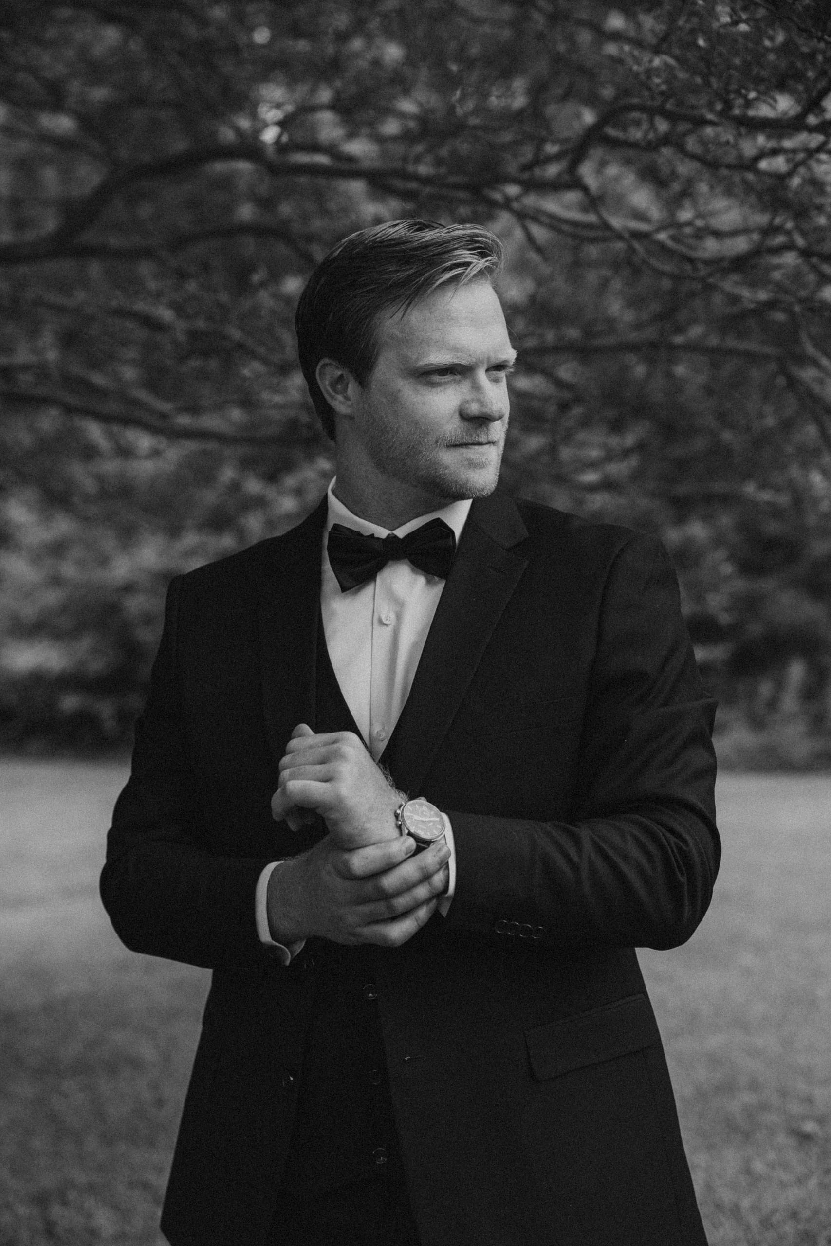 Black and white photo of Andrew in his wedding suit, arranging his buttons and gazing into the distance, creating a timeless and reflective moment.