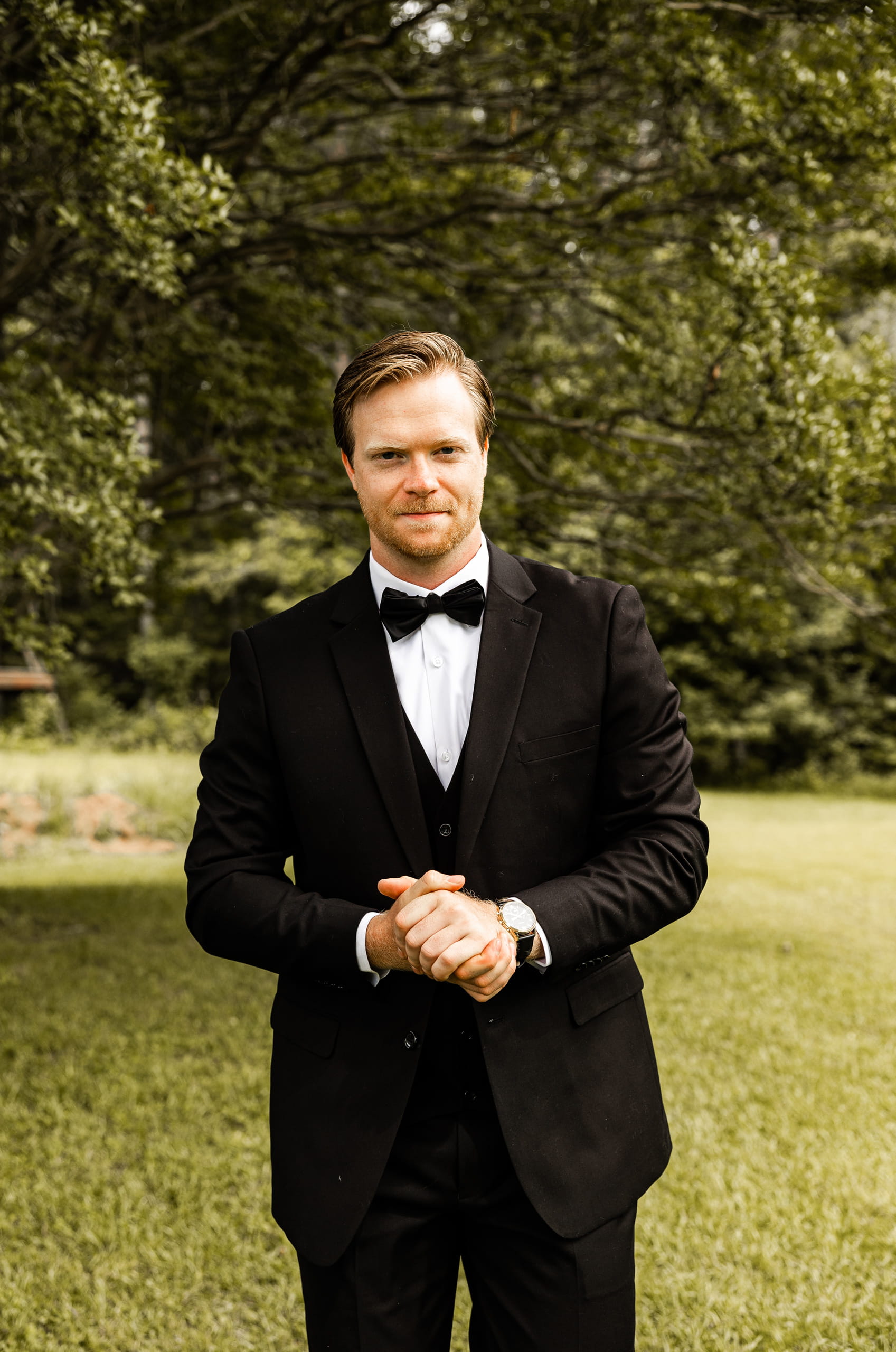 Candid image of Andrew dressed in his wedding suit, standing on the grass in the backyard, surrounded by trees and soft natural light.