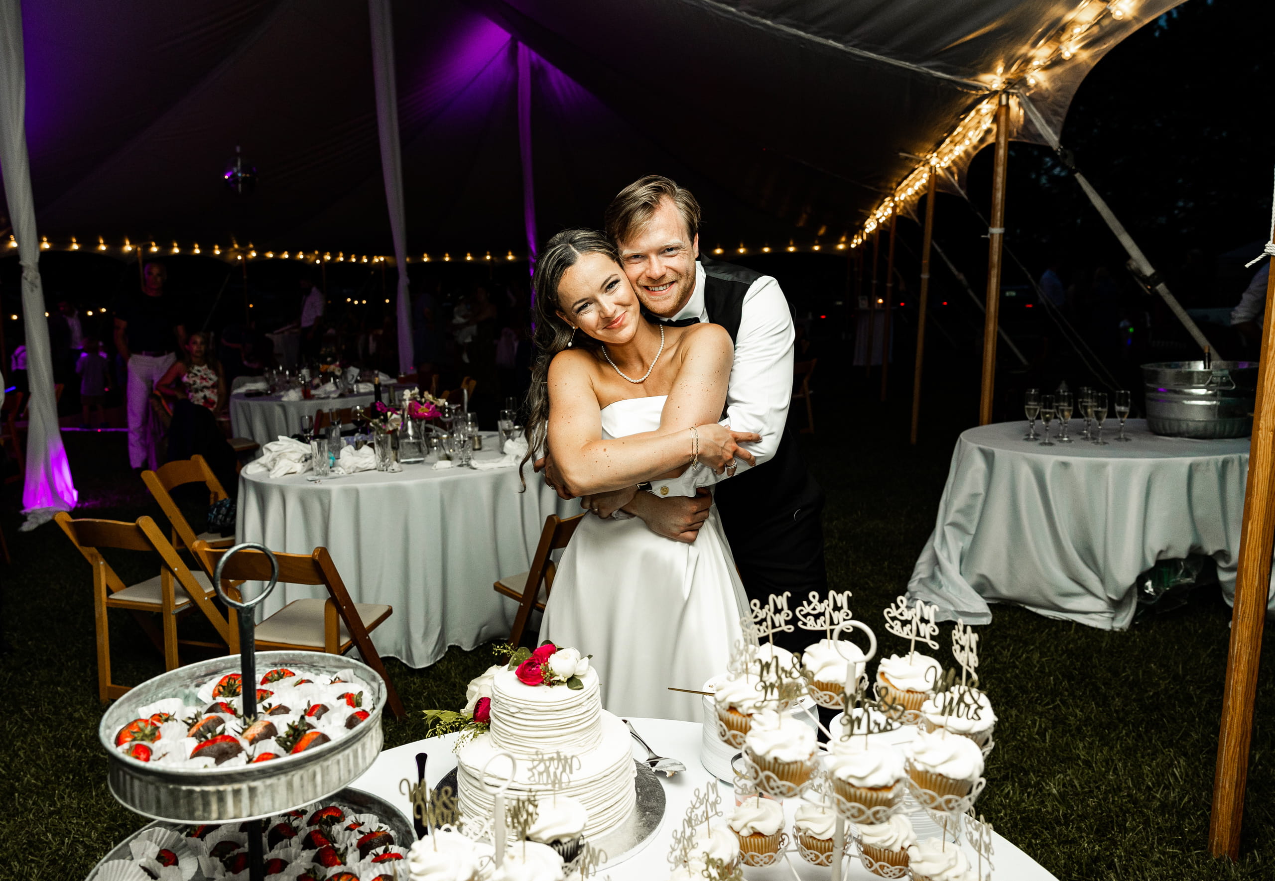 Bride and groom in a warm embrace beside their wedding cake, captured in a romantic and heartfelt moment at the reception.