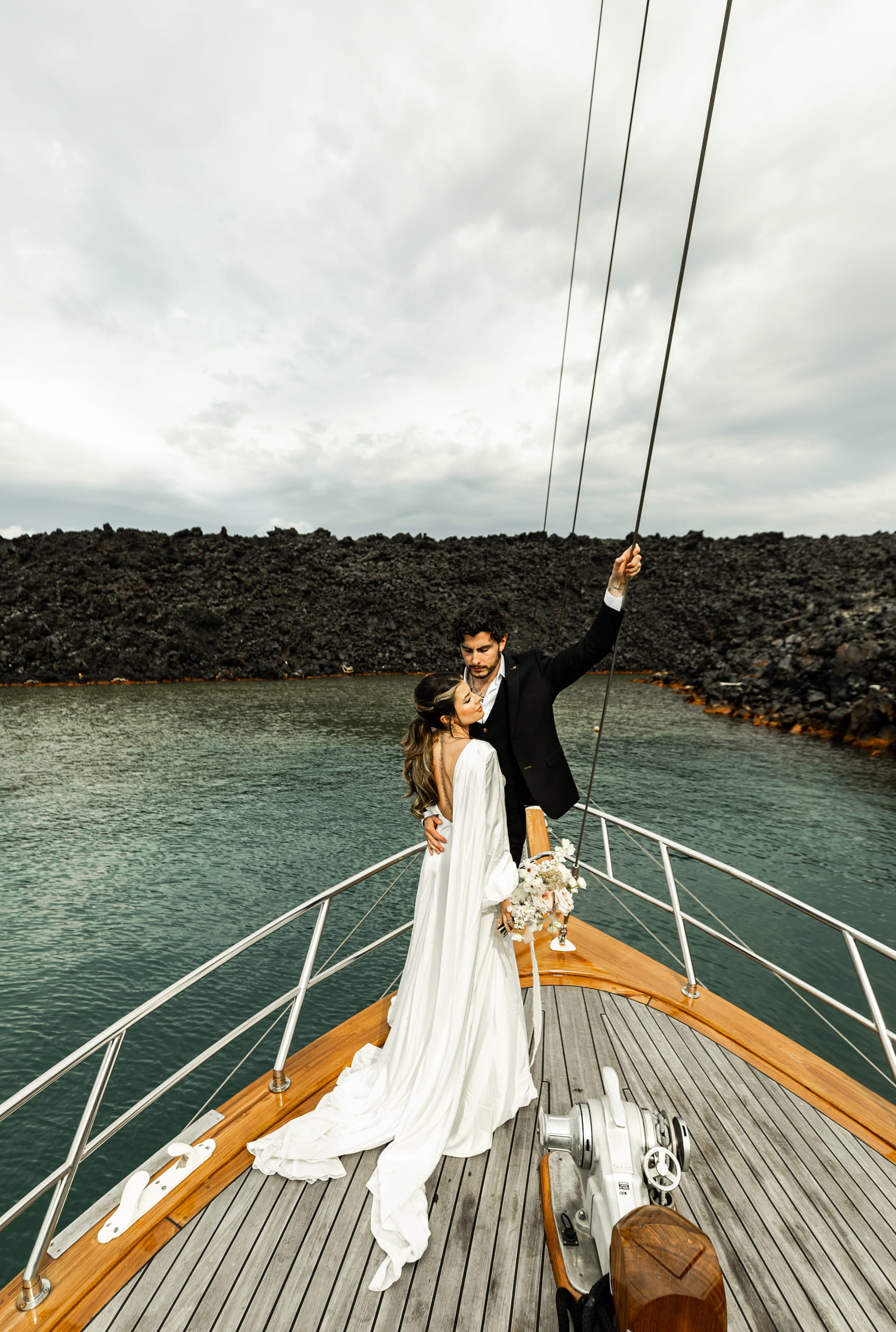 Jeannie and Matty standing on a sailboat in Santorini, sharing a quiet embrace surrounded by volcanic cliffs and blue Aegean waters.