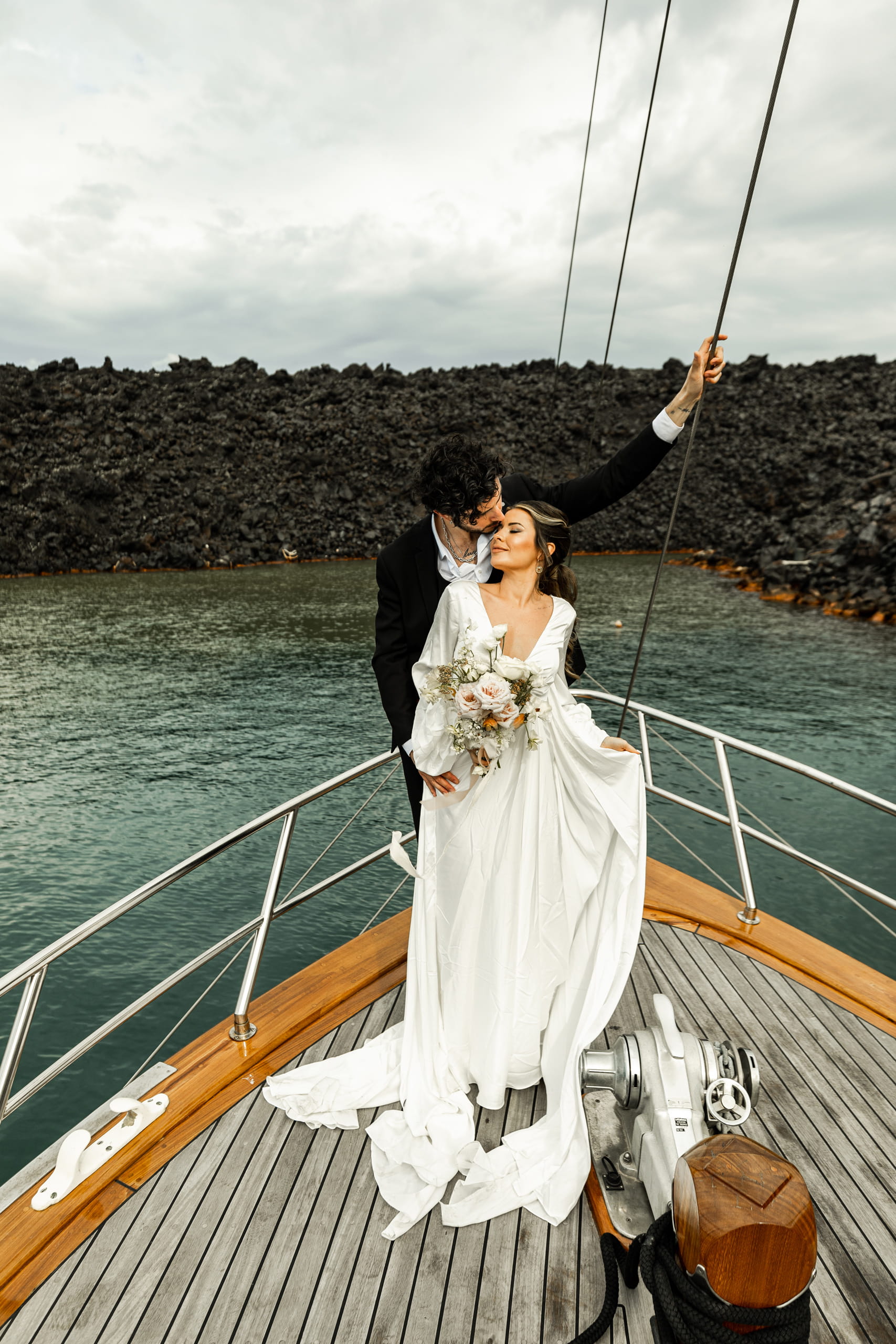 Elegant elopement photo of Jeannie and Matty in front of Santorini’s volcanic cliffs, the sailboat floating gently on the calm Aegean Sea.