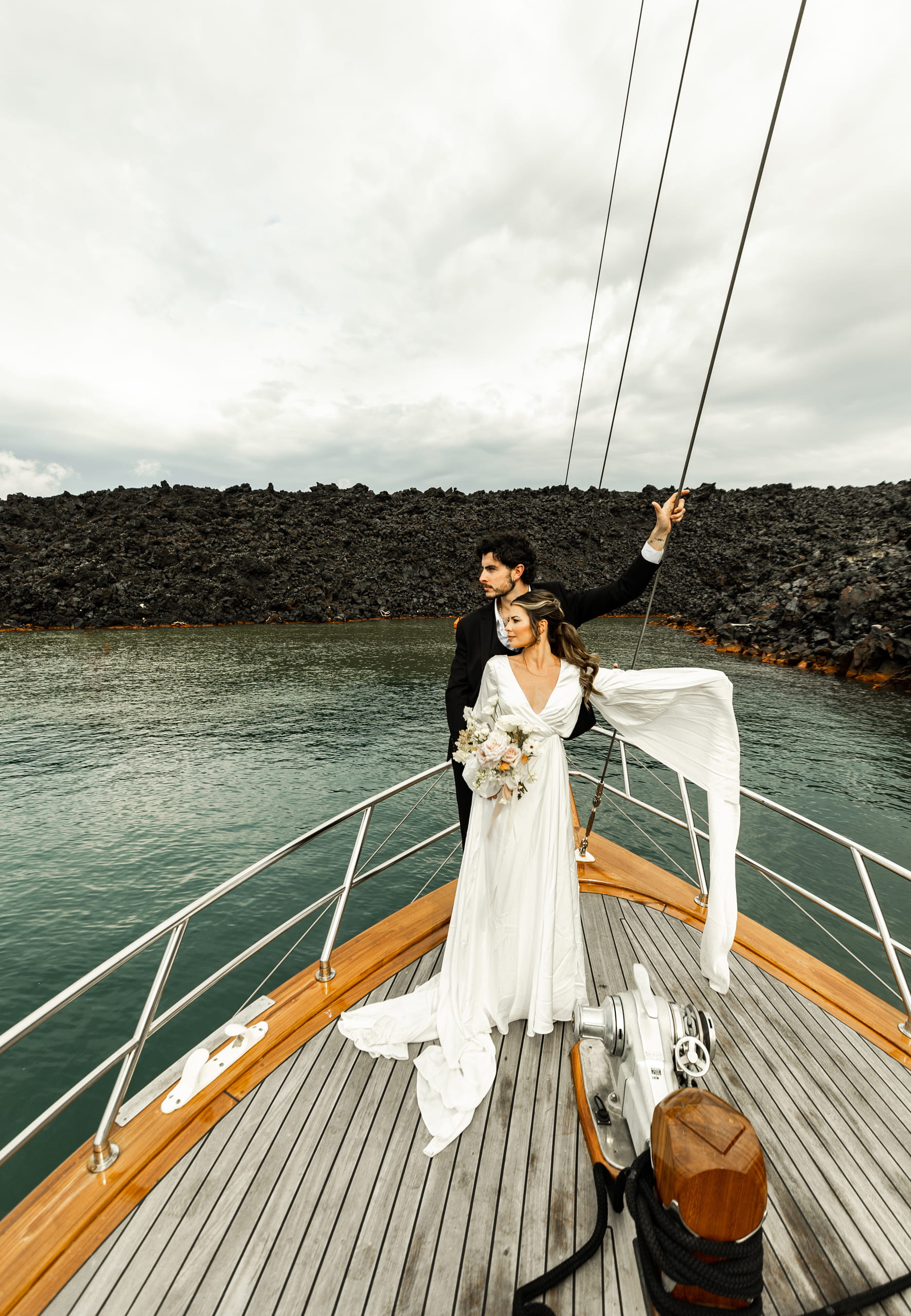 Jeannie holding her floral bouquet close as the sea breeze lifts her veil, with Matty by her side on a sailboat near Santorini’s cliffs.