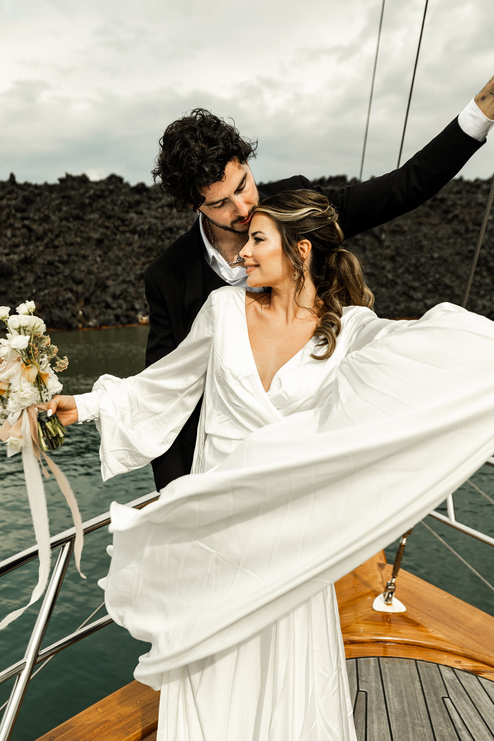 Bride Jeannie laughing beside groom Matty on a boat in Santorini, her bouquet glowing under the golden island light and blue sea horizon.