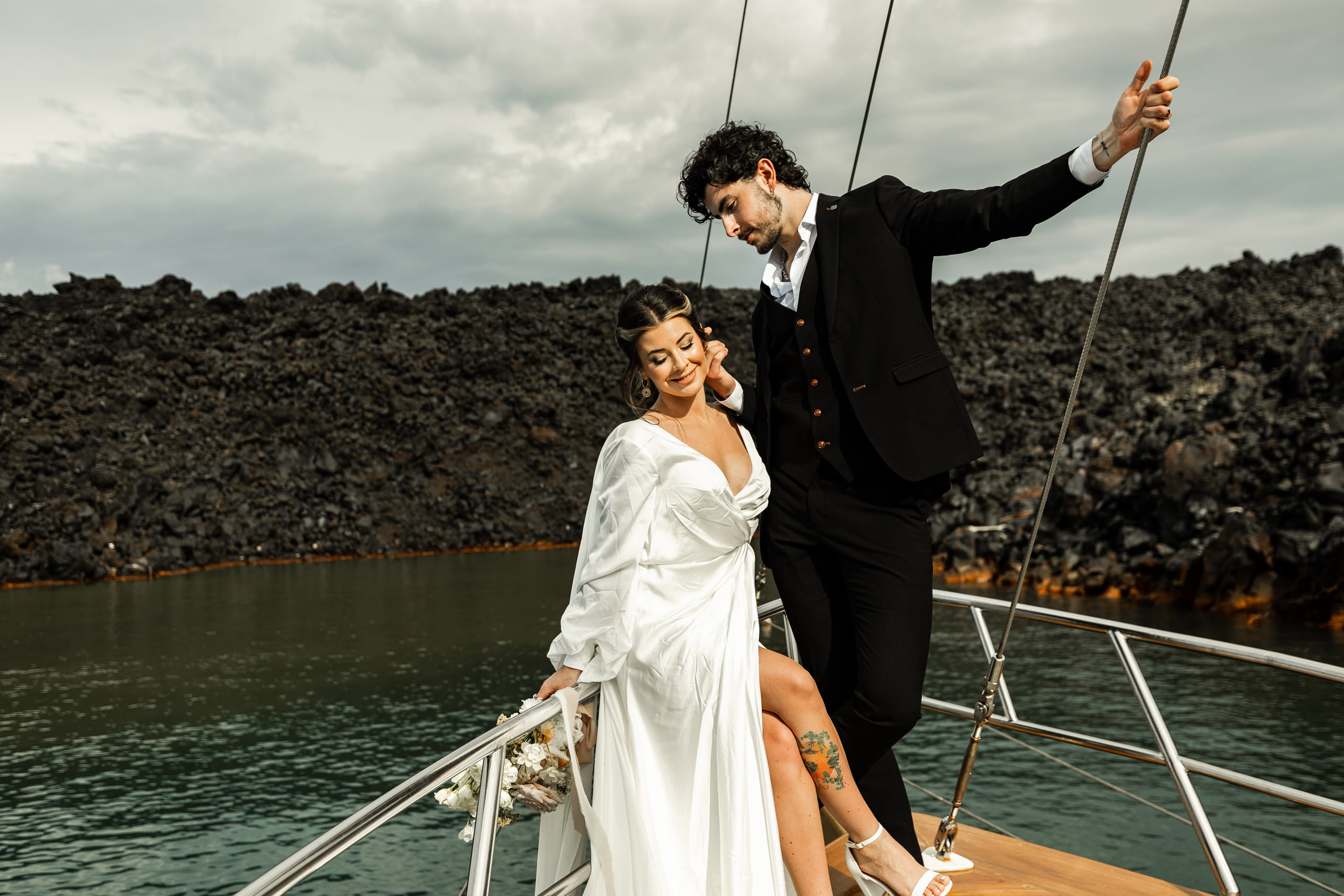 Jeannie and Matty on a boat in Santorini, their silhouettes framed by clouds and distant volcanic rocks.