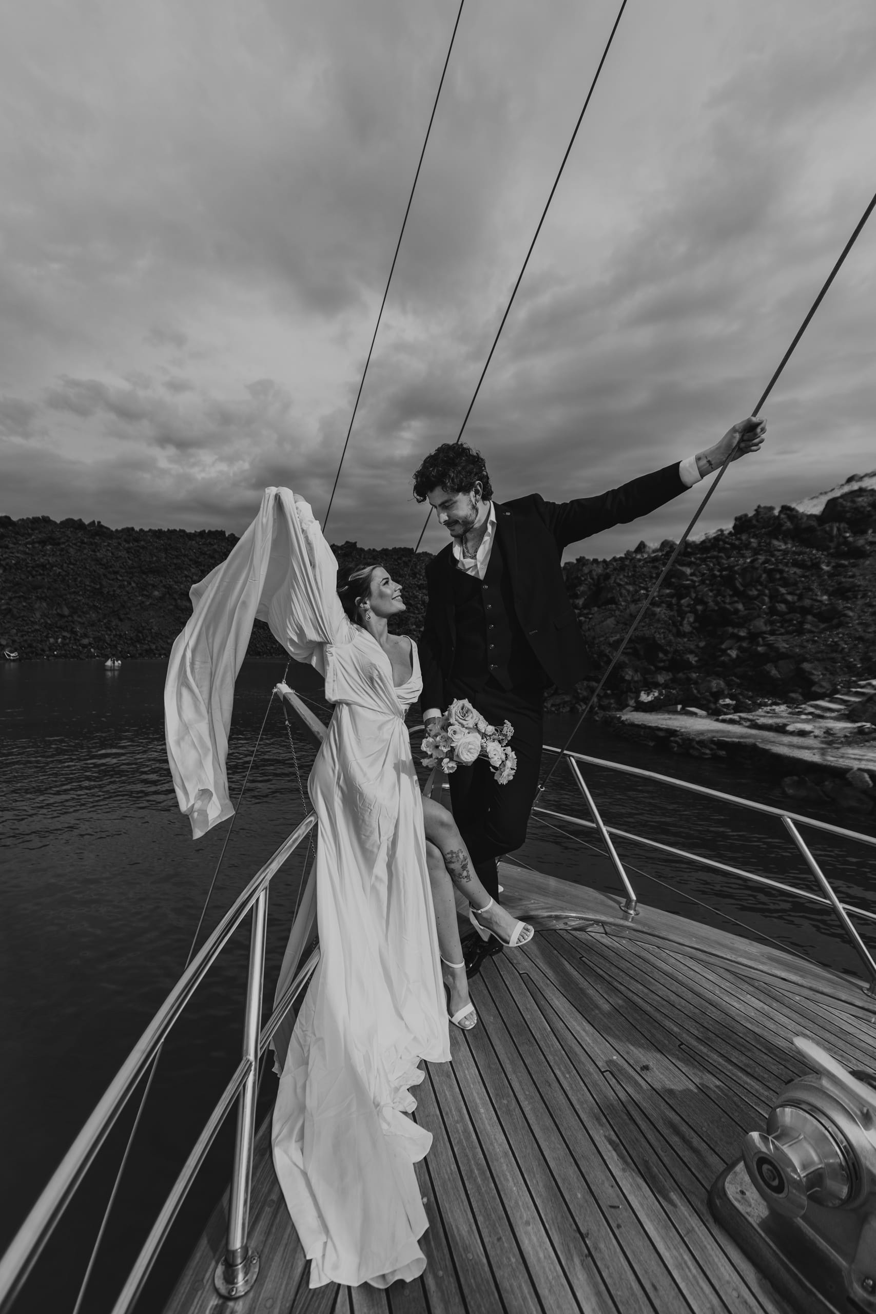 Black and white wedding portrait of Jeannie and Matty in Santorini, the bride showing her veil and bouquet as the groom looks lovingly at her.