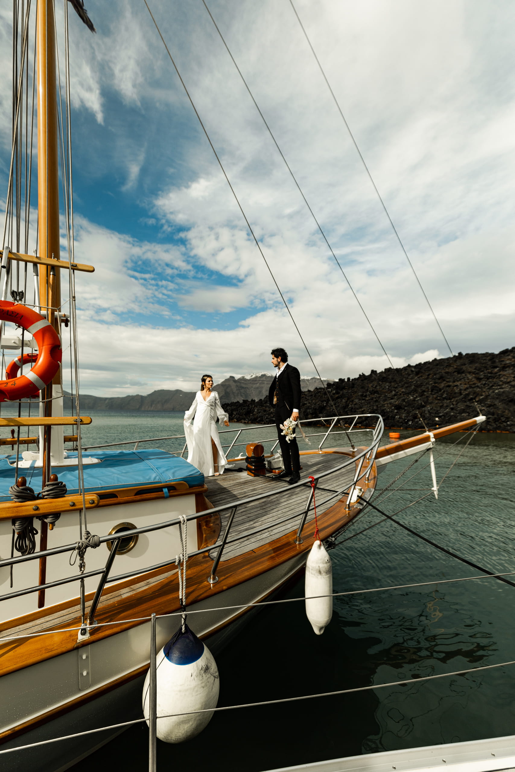 Jeannie and Matty approaching each other on a sailboat in Santorini, with the blue and white sky reflecting over the Aegean Sea.