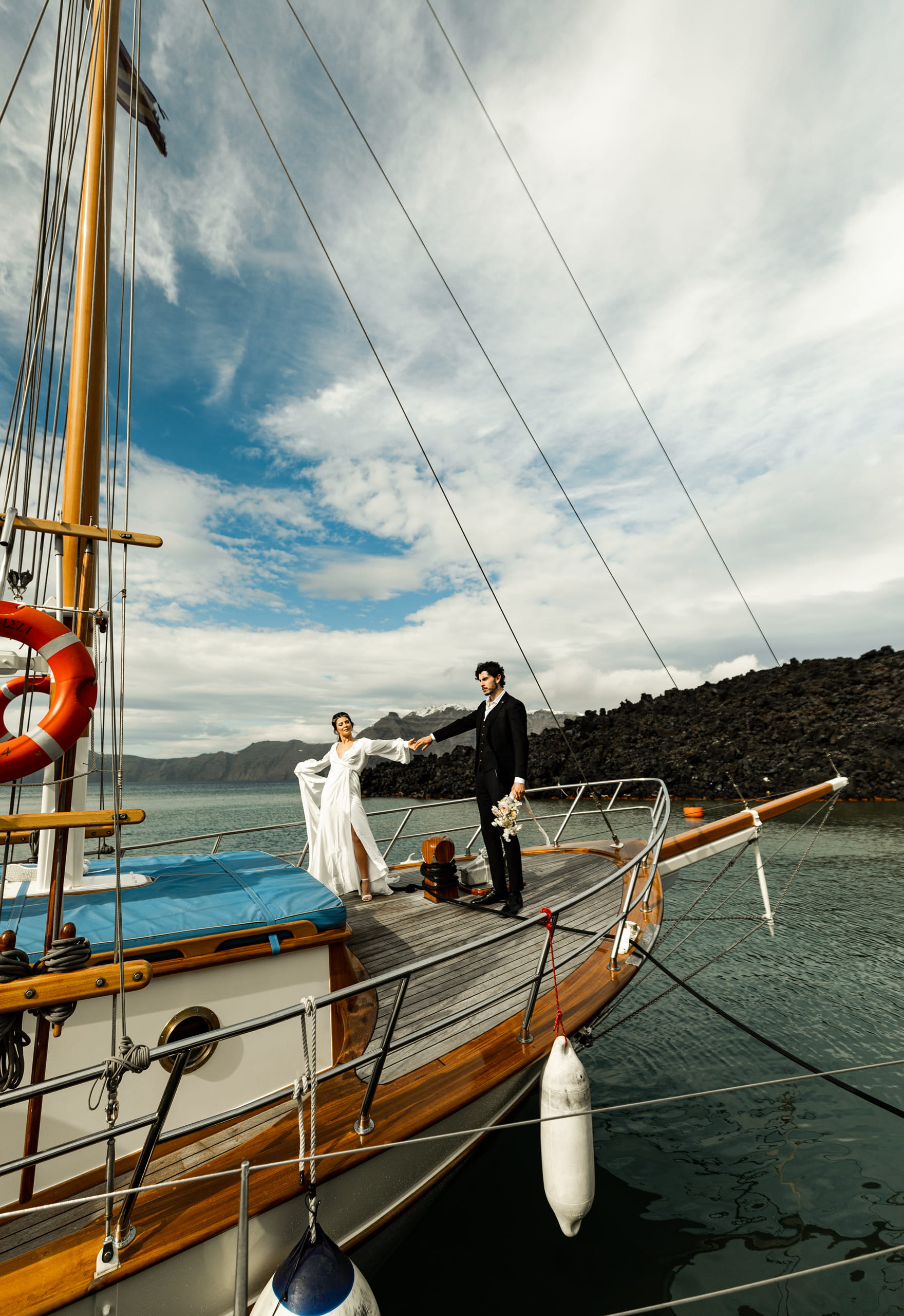 Bride and groom Jeannie and Matty holding hands on a sailboat in Santorini, surrounded by bright blue skies and soft white clouds.