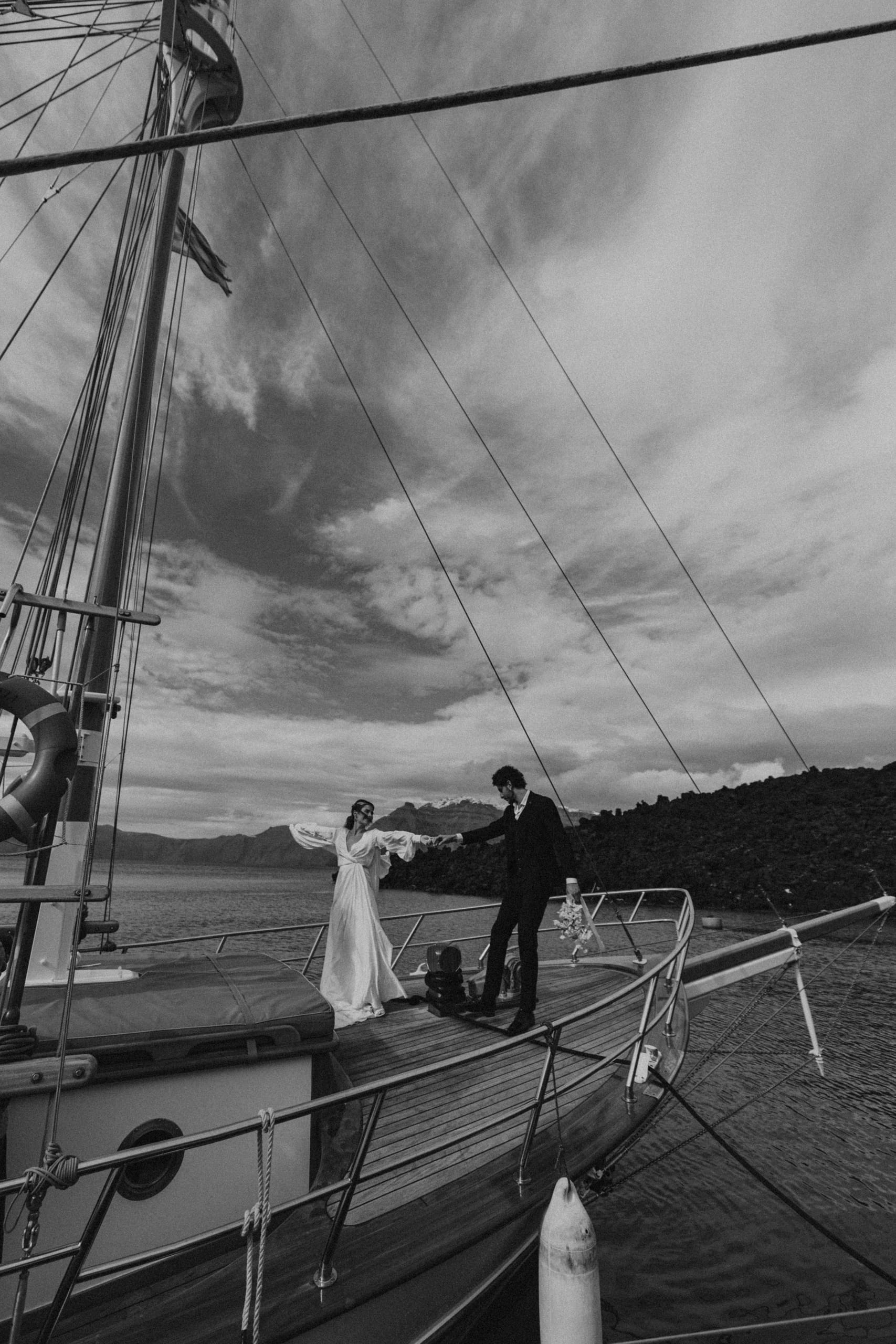 Black and white wedding portrait of Jeannie and Matty holding hands, framed by the splendour of the Santorini sky and the spirit of love.