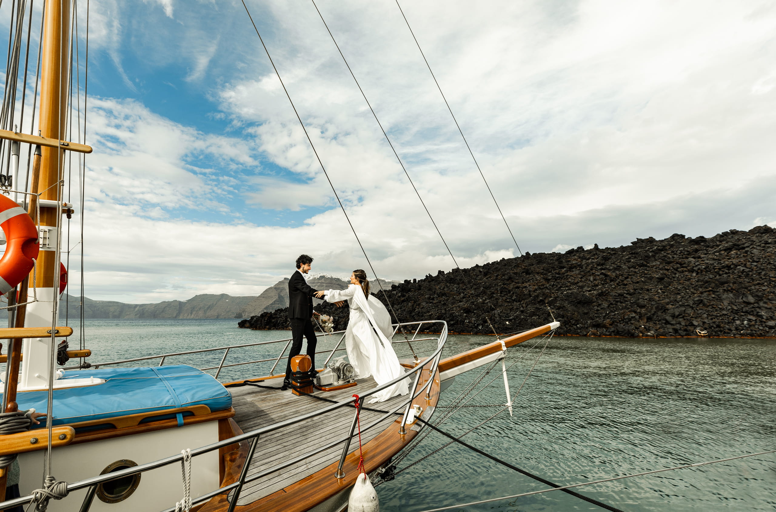 Jeannie reaching out to offer her hand to Matty aboard their wedding sailboat in Santorini, under a radiant blue and white sky.