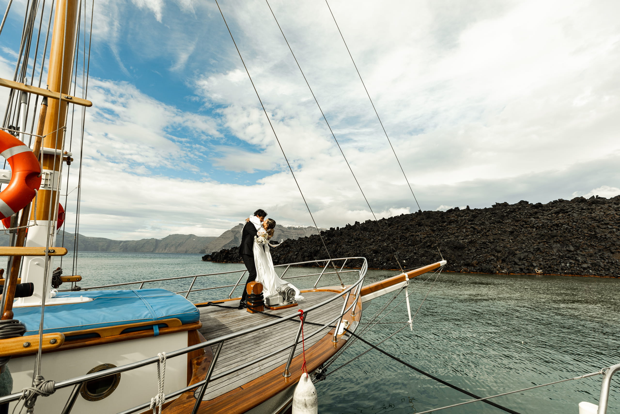 Romantic moment of Jeannie and Matty embracing at the bow of their sailboat in Santorini, surrounded by the calm Aegean Sea.