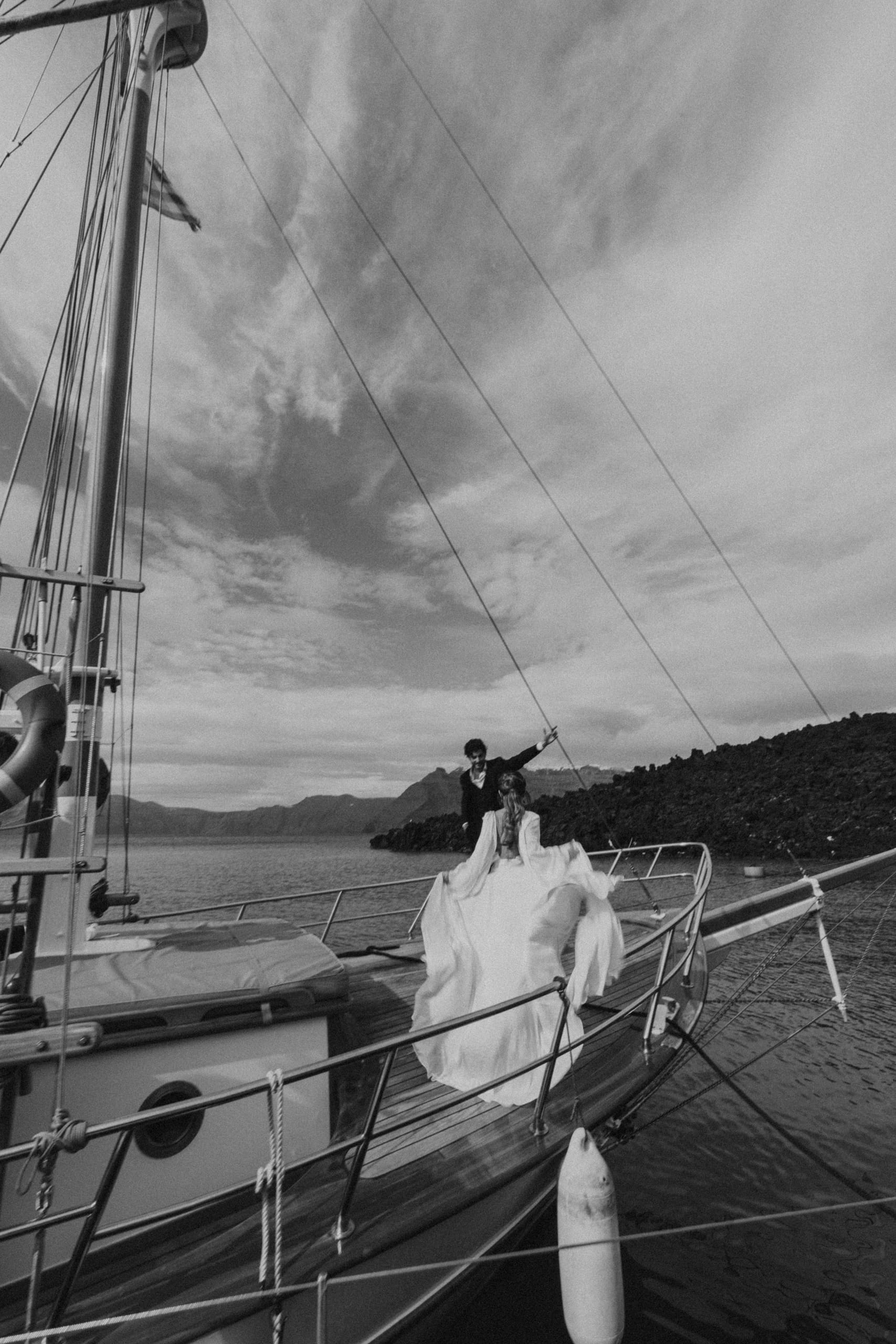Artistic black and white image of the bride opening her arms toward the groom on a sailboat, surrounded by the serene beauty of their Santorini elopement.