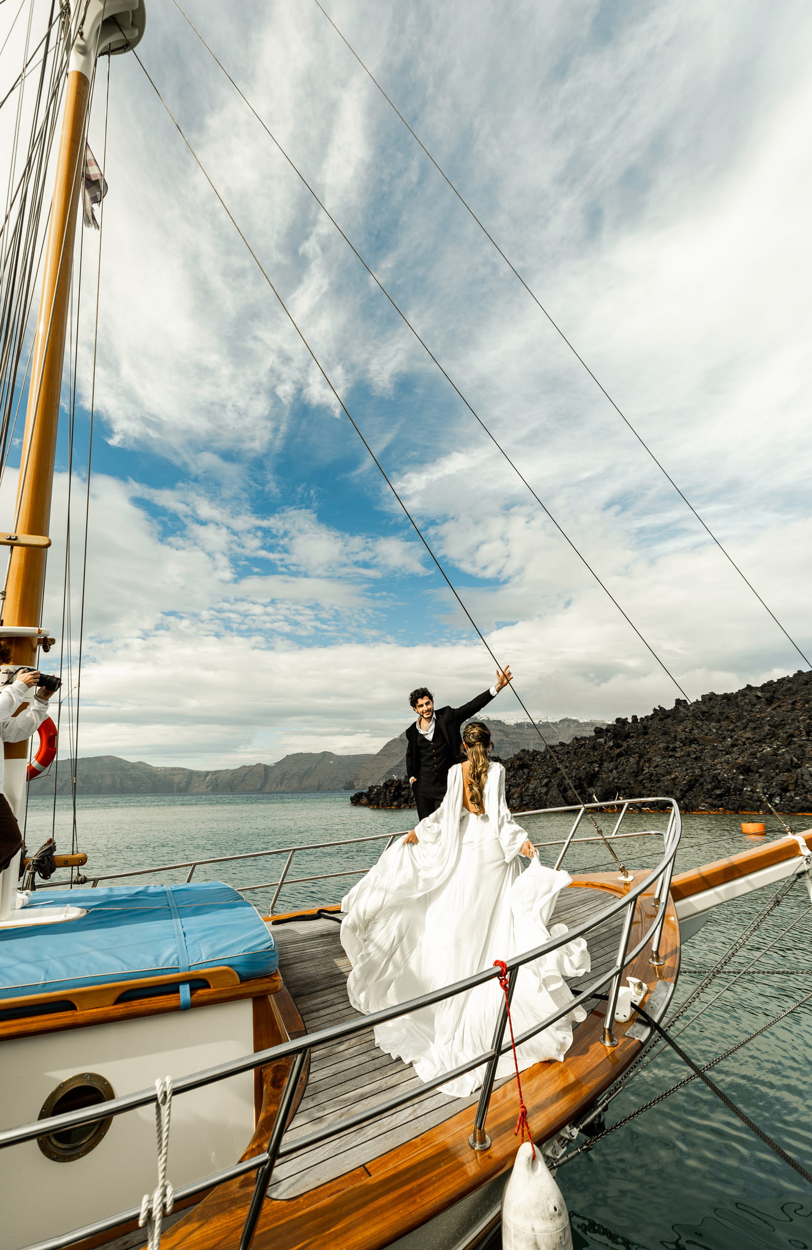 Bride Jeannie holding her dress as she steps forward to meet Matty on their sailboat in Santorini, surrounded by the blue Aegean Sea and volcanic cliffs.