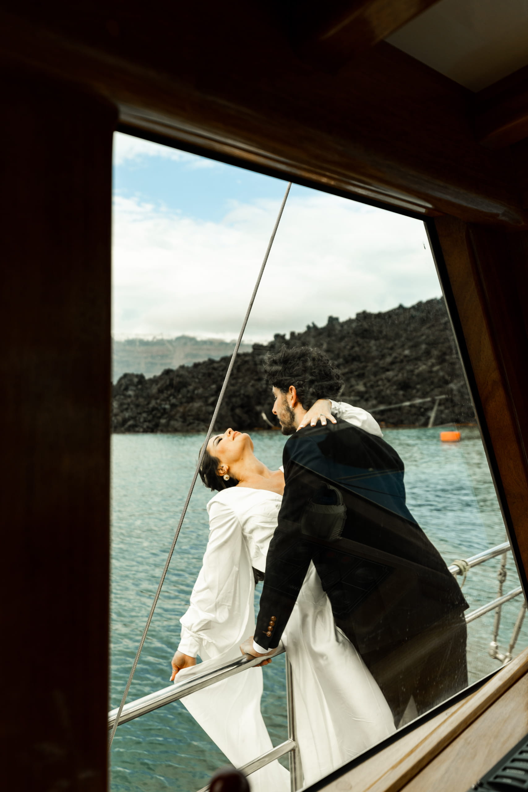 Jeannie lying back on the side of the sailboat looking toward the sky as Matty holds her with his arm around her neck, capturing a tender Santorini elopement moment.