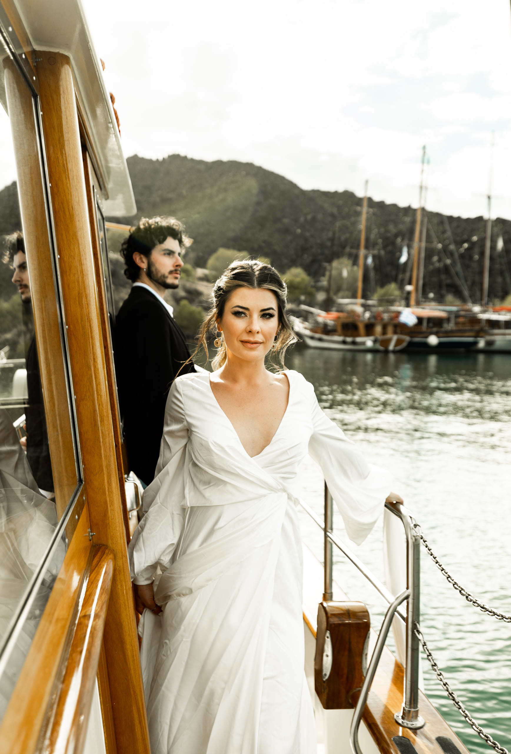 Jeannie and Matty on their sailboat approaching the Santorini harbour, the groom leaning on the side looking at the horizon while the bride smiles toward the camera showing her wedding dress.