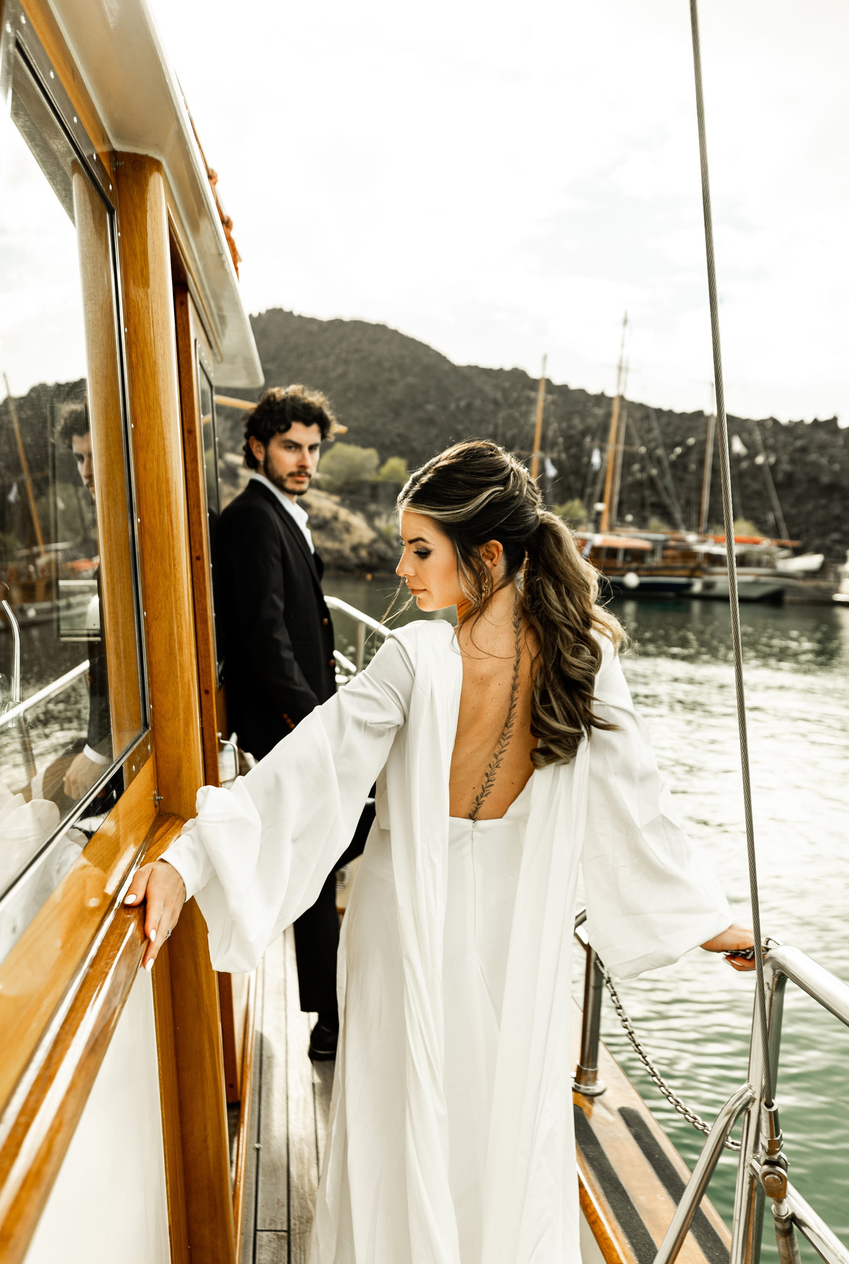 Bride Jeannie facing away from the camera showing her gown as Matty looks lovingly at her on the sailboat near Santorini’s volcanic cliffs.