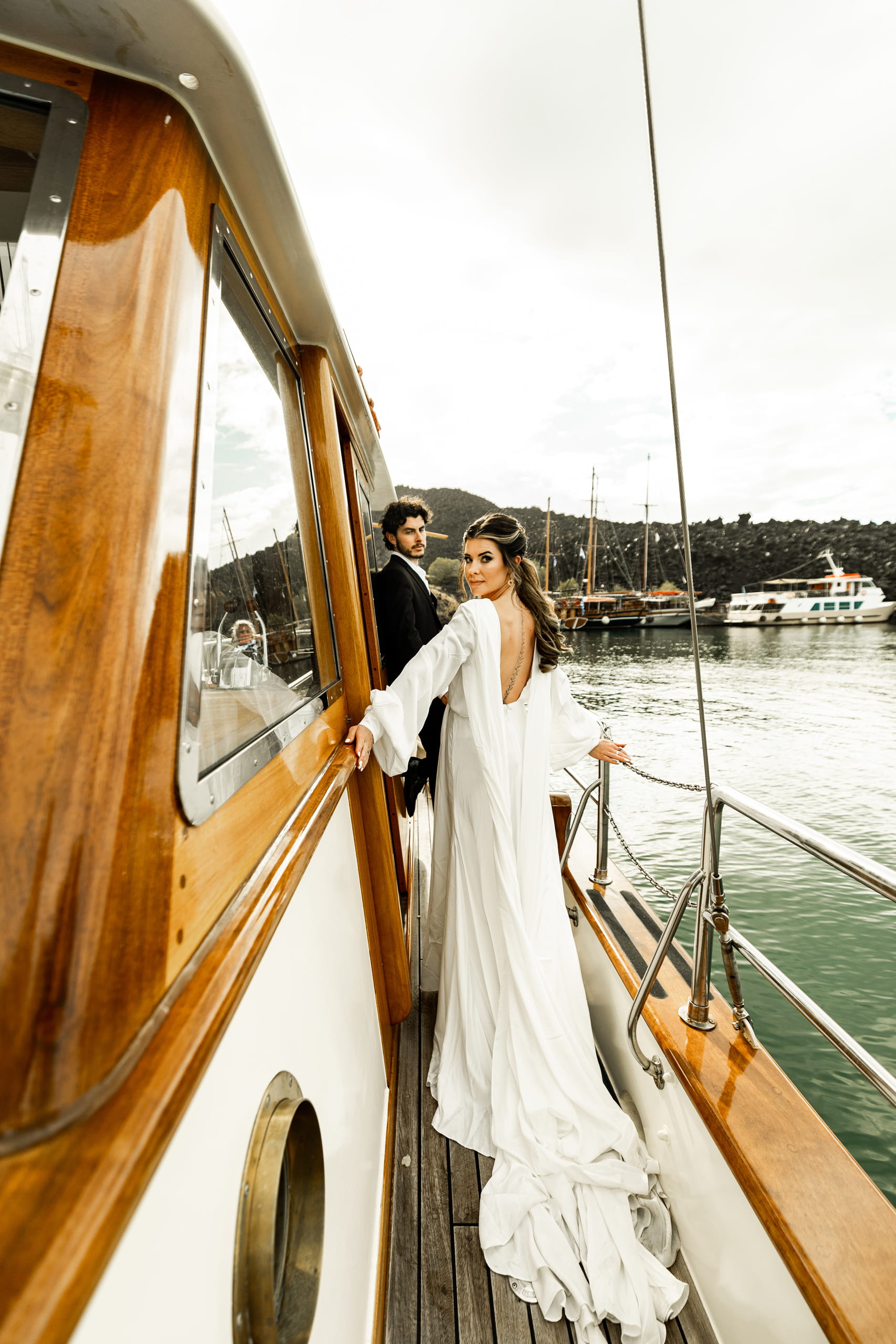 Jeannie and Matty standing together on the sailboat near Santorini, captured from afar as they pose gracefully against the Aegean horizon.