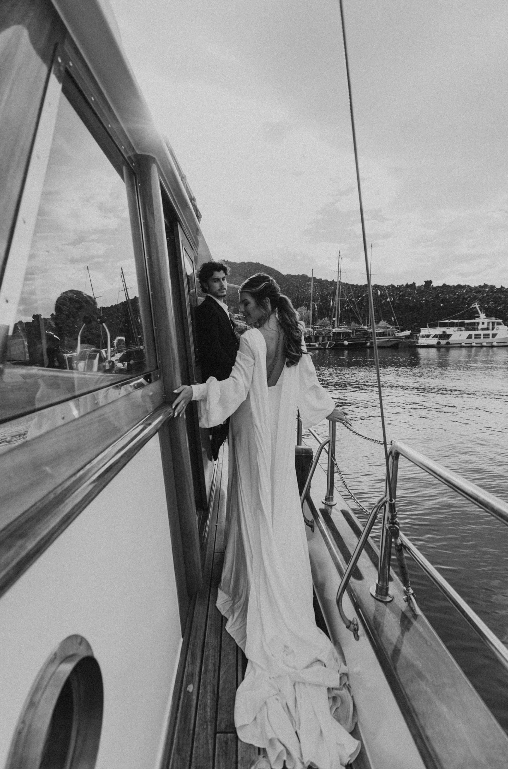 Black and white photo of Jeannie turned away from the camera showing her wedding dress as Matty gazes at her on their sailboat in Santorini.