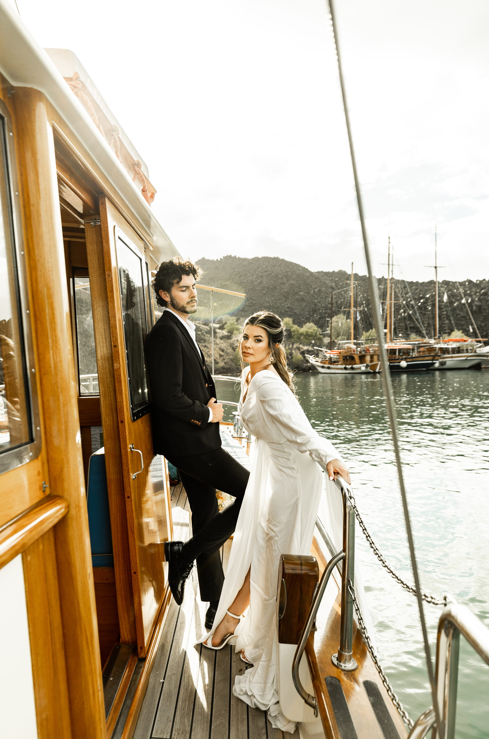 Santorini elopement photo of Jeannie and Matty on their sailboat nearing the harbour, the bride posing gracefully from the side with her gown flowing in the breeze.