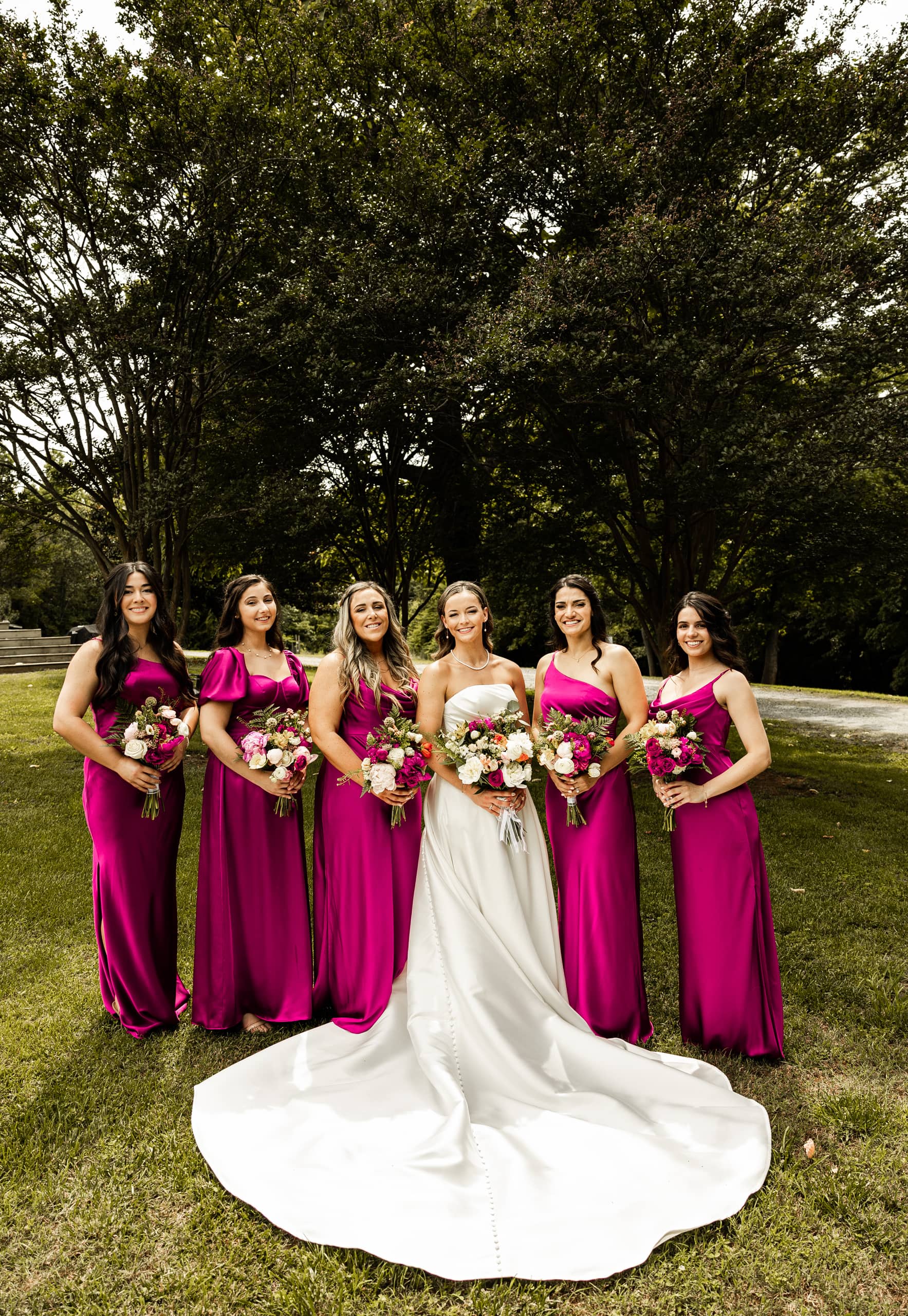 Bride Alyssa in her white wedding dress standing with her bridesmaids in magenta dresses, all holding flower bouquets in the backyard garden.