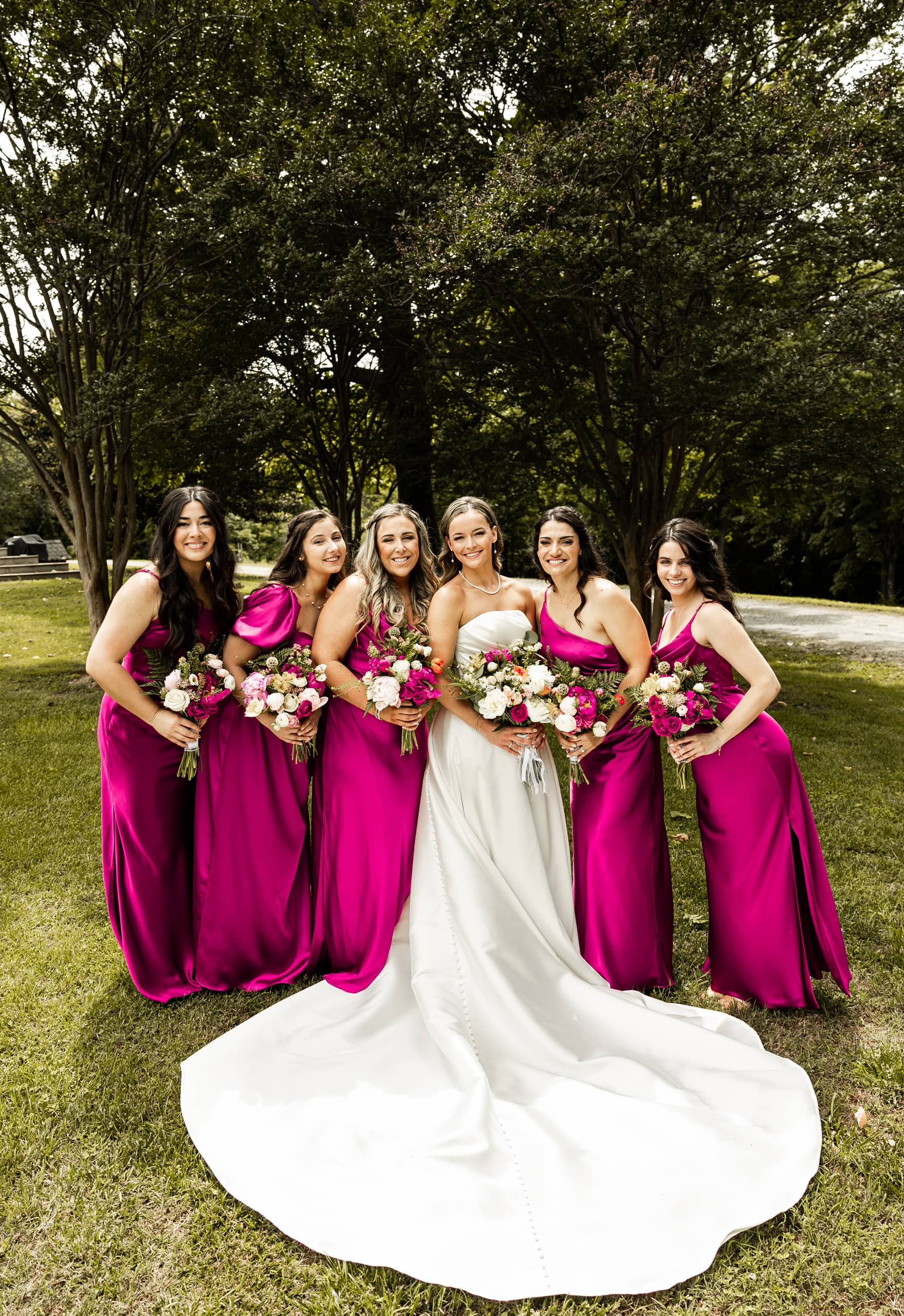 Joyful moment of the bride and bridesmaids dressed in magenta gowns with floral bouquets, gathered beside Alyssa in her wedding dress in the backyard.