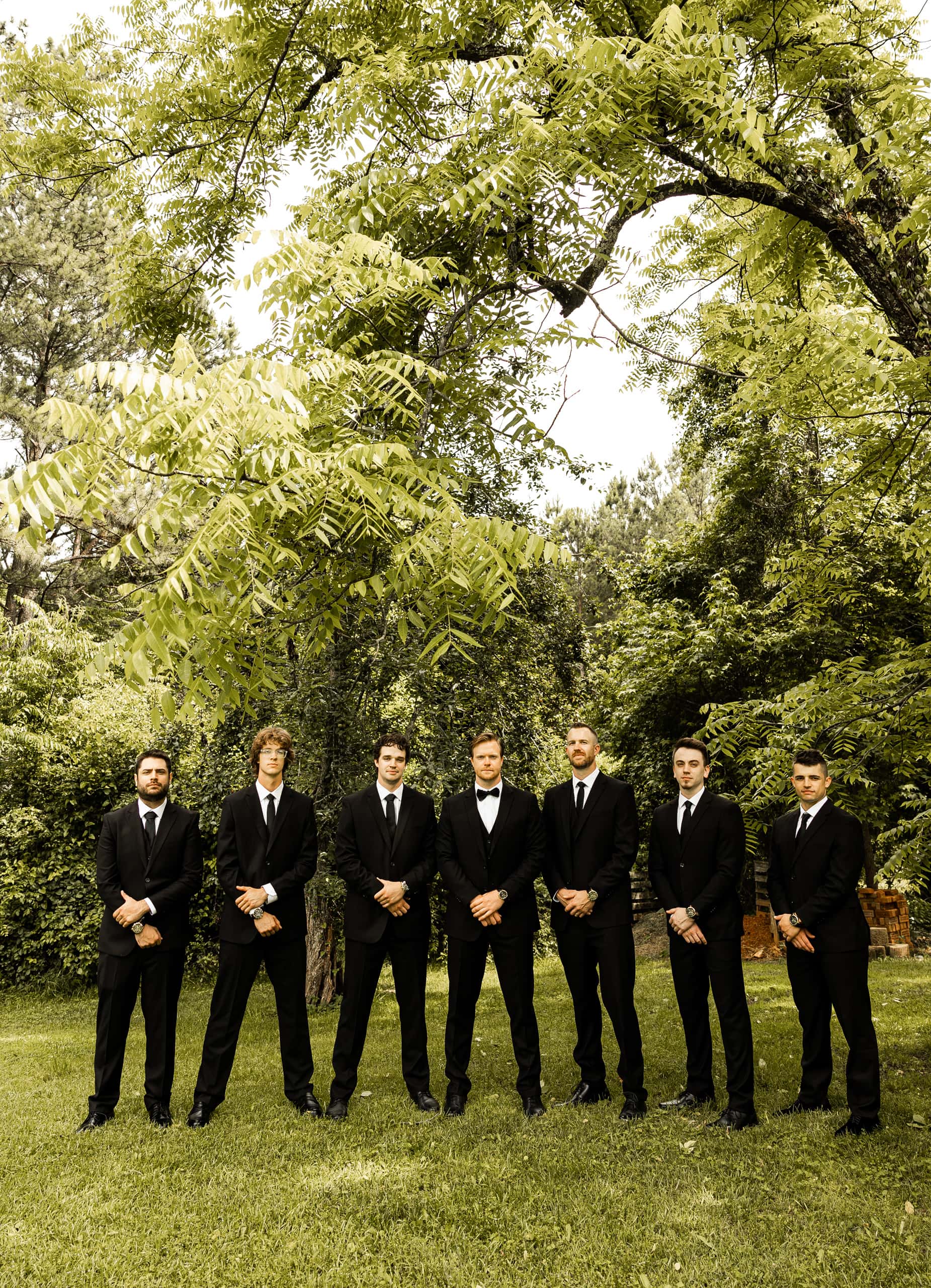 Andrew and his groomsmen standing side by side in the backyard with their hands crossed in front, captured in a classic and composed wedding portrait.