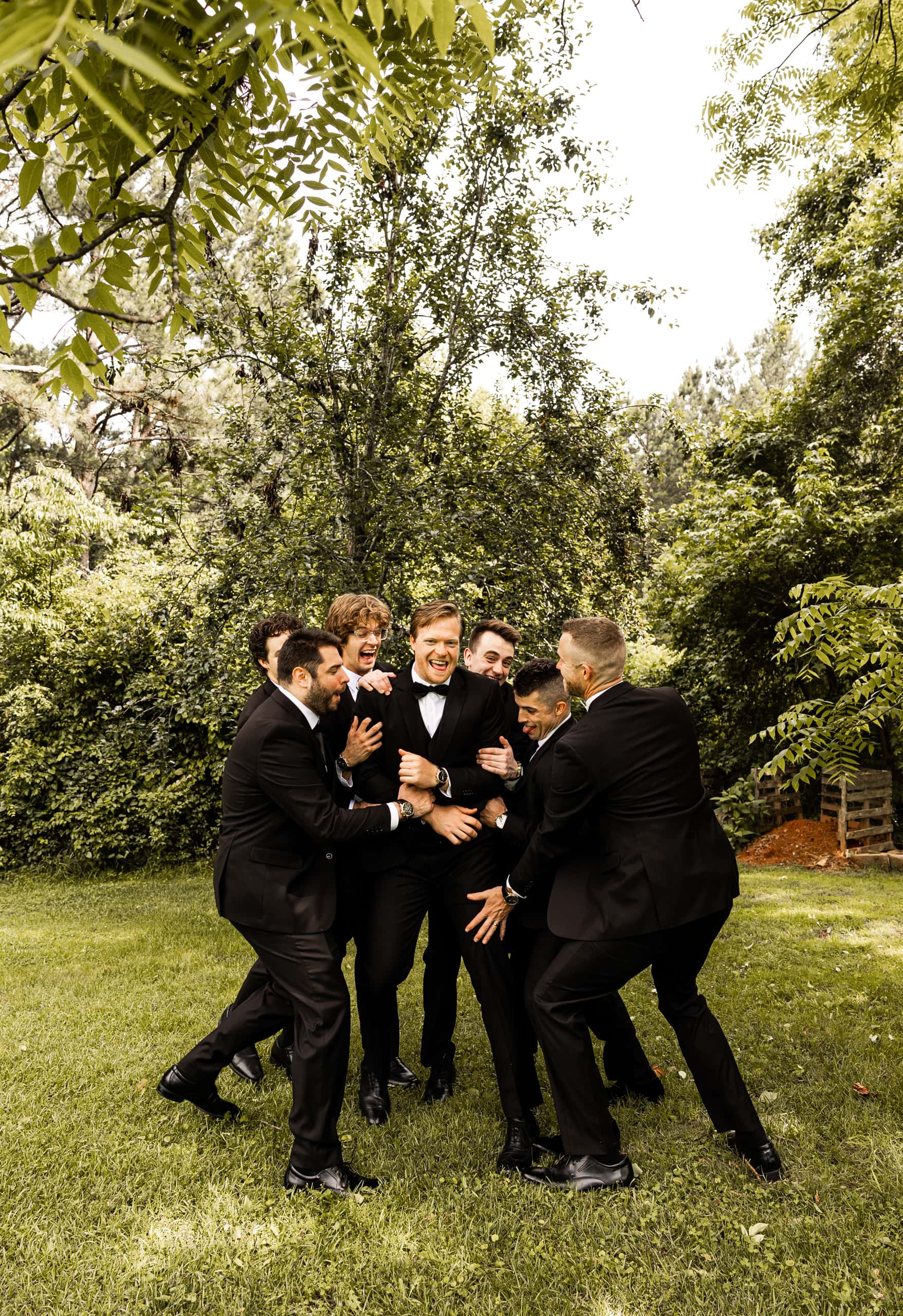Groomsmen laughing as they try to lift Andrew in his wedding suit during a playful moment in the backyard before the ceremony.