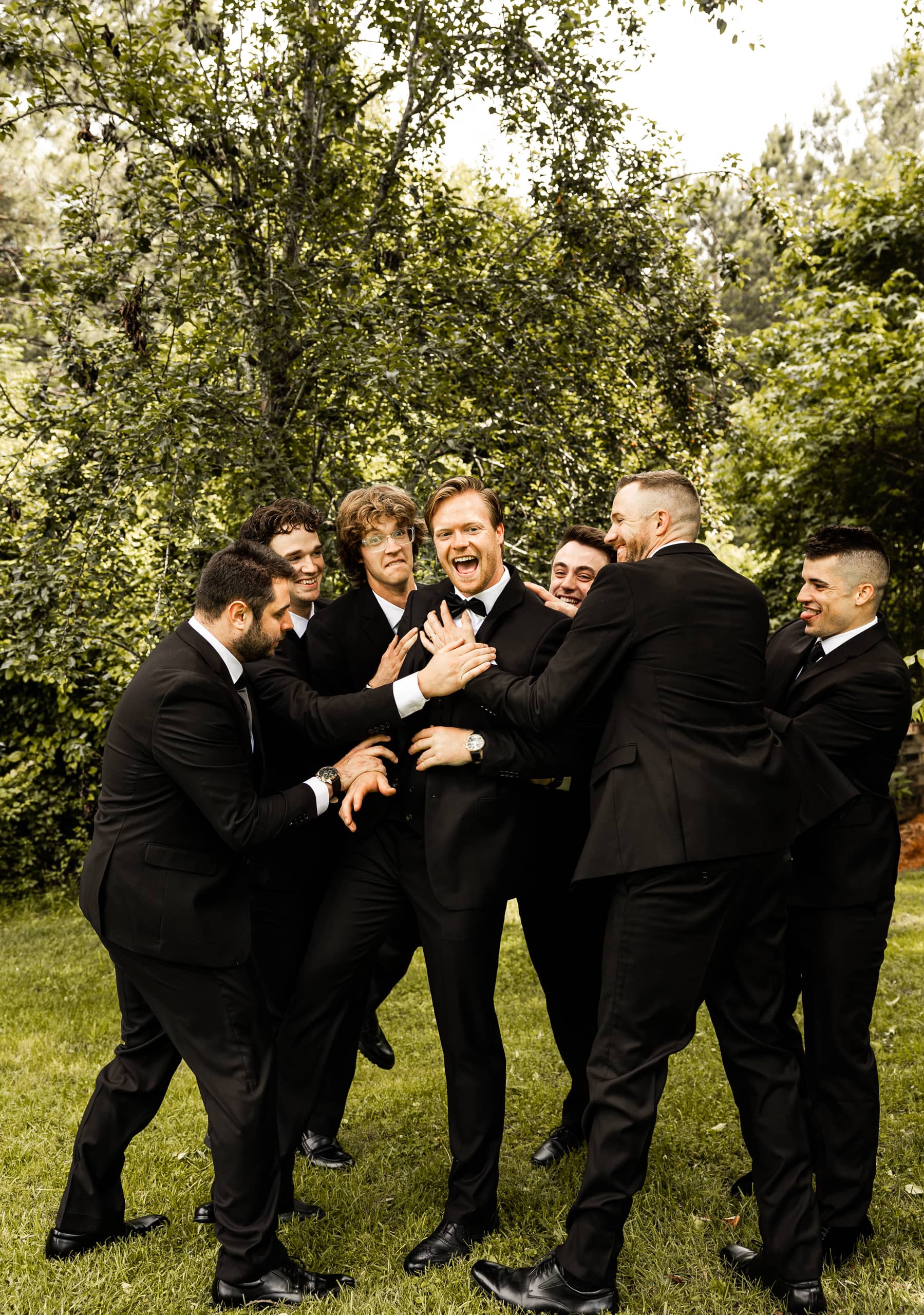 Candid photo of Andrew and his groomsmen enjoying a joyful moment outdoors as the group attempts to lift him up amid laughter and celebration.